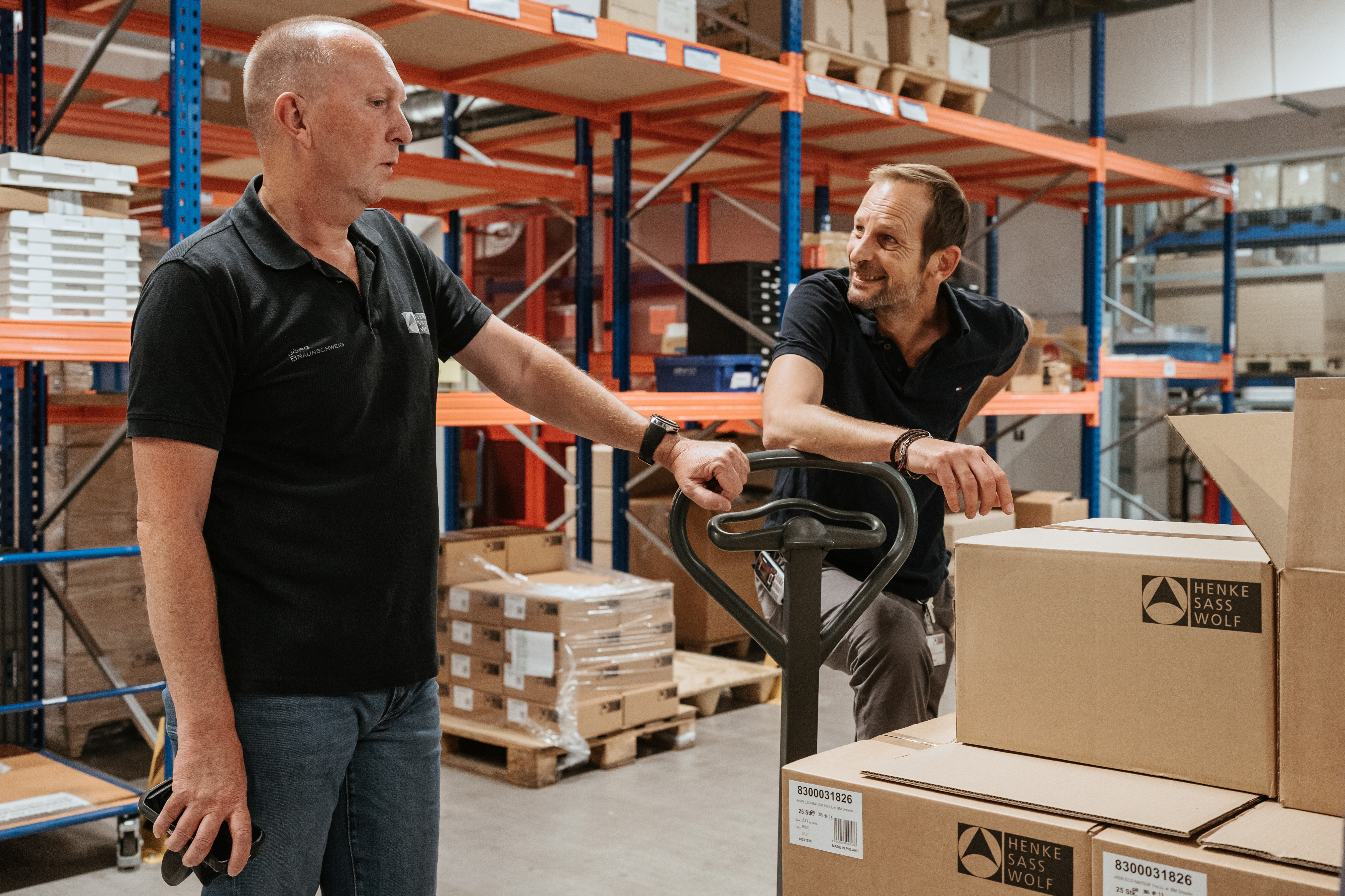 Zwei%20Henke-Sass%2C%20Wolf%20Lagermitarbeiter%20im%20Gespr%E4ch%2C%20einer%20h%E4lt%20einen%20Handscanner%2C%20umgeben%20von%20beschrifteten%20Versandkartons%20und%20Hochregalen